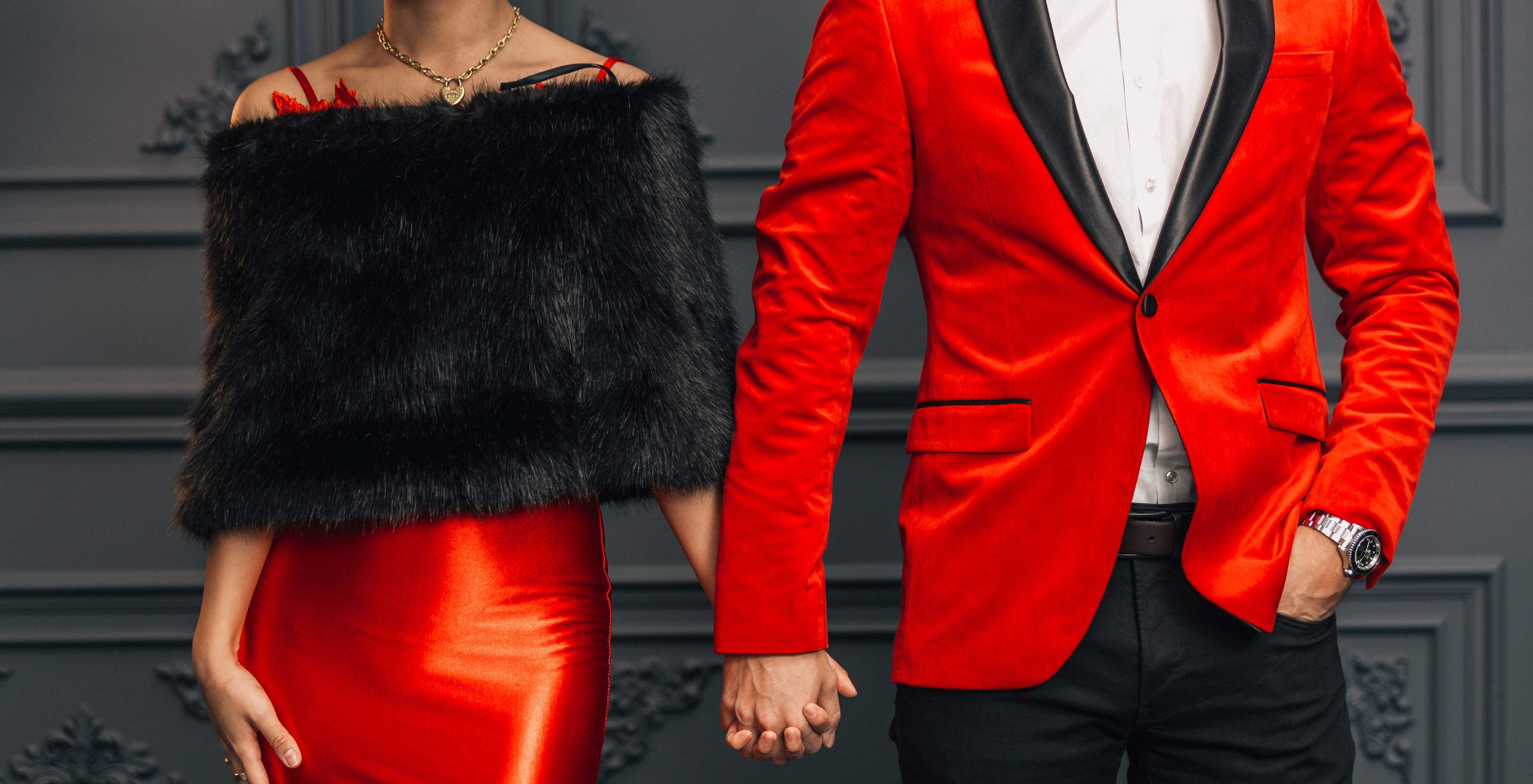 A couple in formal evening wear holding hands. The woman wears a bright red satin off-the-shoulder dress with a black faux-fur bolero.